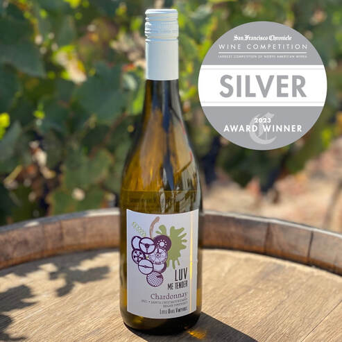 bottle of LUV's Chardonnay on a wine barrel with vines in the background and silver award badge over image